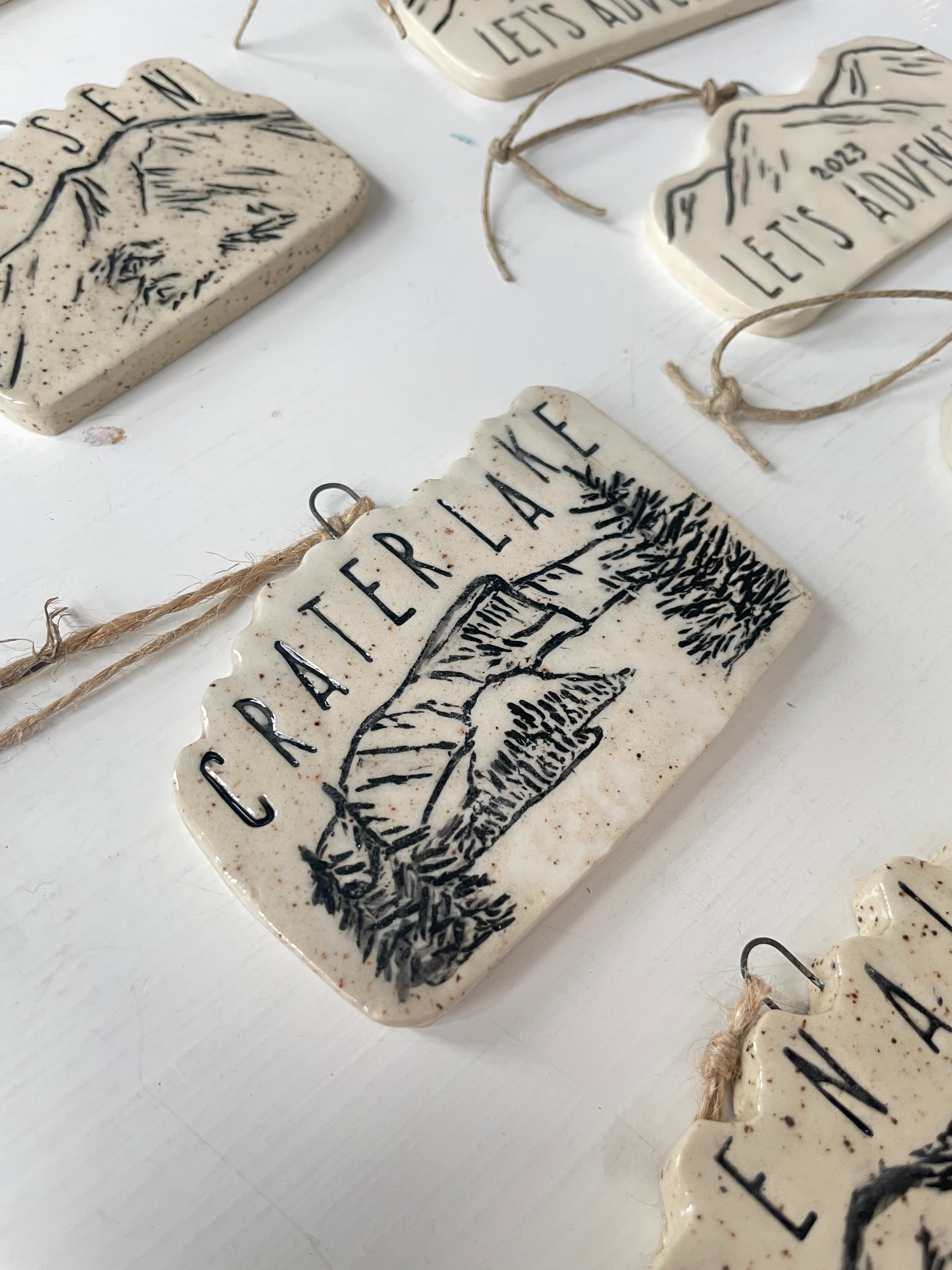 Hand-Carved Ceramic Ornaments. Black Ink designs of mountains and national parks. Shown is a design of Crater Lake.